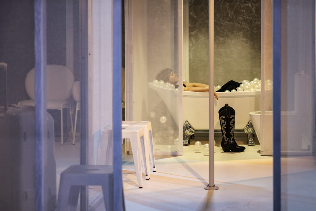 Zdjęcie ze spektaklu Tramwaj zwany pożądaniem. Na pierwszym planie jest rura do tańczenia i przezroczyste ściany. Z tyłu młoda kobieta z krótkimi włosami leży w ubraniu w wannie. Wanna jest wypełniona drobnymi piłkami. Obok stoją buty kowbojki.