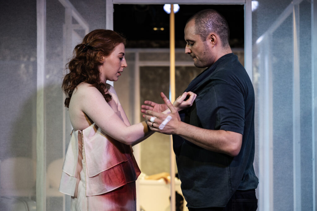 Zdjęcie ze spektaklu Tramwaj zwany pożądaniem. Młoda kobieta z kręconymi włosami i młody mężczyzna stoją naprzeciwko siebie. Kobieta trzyma mężczyznę za koszulkę. Mężczyzna dotyka jej rąk. Mężczyzna na dłoni ma obrączkę i dwa plastry.