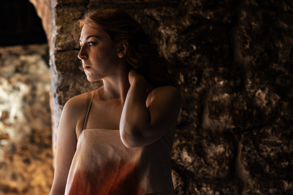 Zdjęcie ze spektaklu Tramwaj zwany pożądaniem. Młoda kobieta z kręconymi włosami opiera się o ścianę z kamieni. Jest poważna, patrzy w bok, jedną rękę trzyma na szyi.