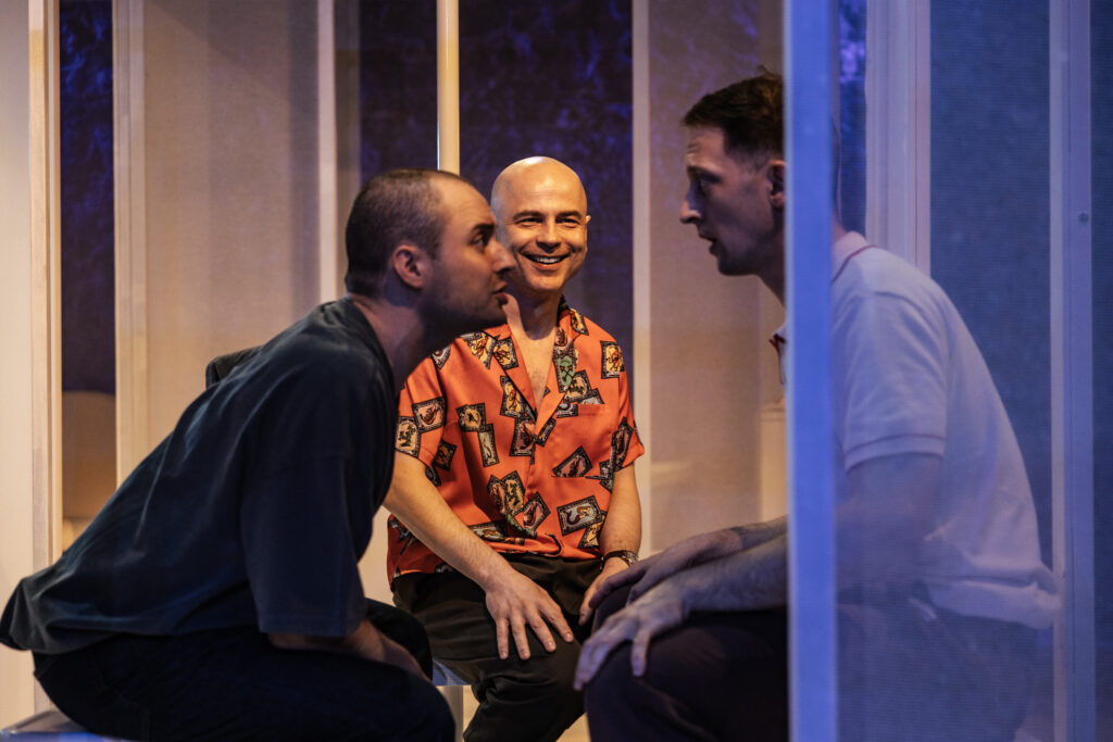 Zdjęcie ze spektaklu Tramwaj zwany pożądaniem. Trzej mężczyźni siedzi naprzeciwko siebie, rozmawiają. W tle przeźroczyste ściany.