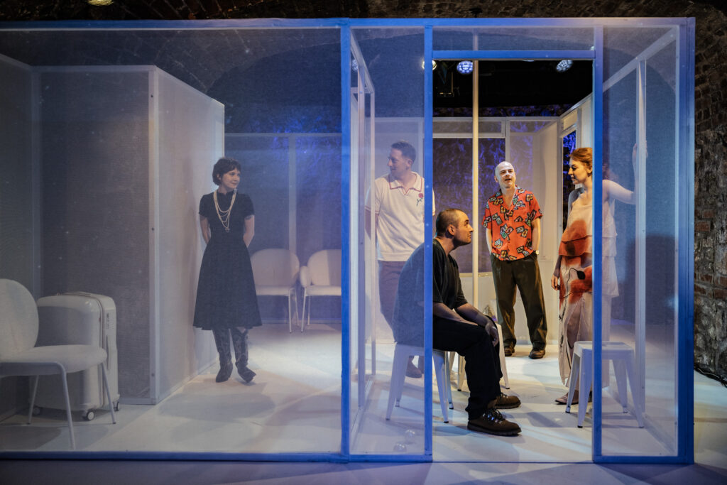 Zdjęcie ze spektaklu Tramwaj zwany pożądaniem. 5 osób w mieszkaniu z przezroczystymi ścianami. Z prawej strony młody mężczyzna siedzi na krześle obok stoi kobieta z kręconymi włosami. Za nimi stoją dwaj mężczyźni. Z lewej strony stoi druga kobieta.