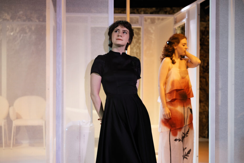 Zdjęcie ze spektaklu Tramwaj zwany pożądaniem. Dwie młode kobiety. Pierwsza ma krótkie włosy i ciemną sukienkę opiera się o przezroczystą ścianę. Druga stoi za nią, ma kręcone włosy i jasną sukienkę. W tle minimalistyczne mieszkanie, wanna i krzesła.