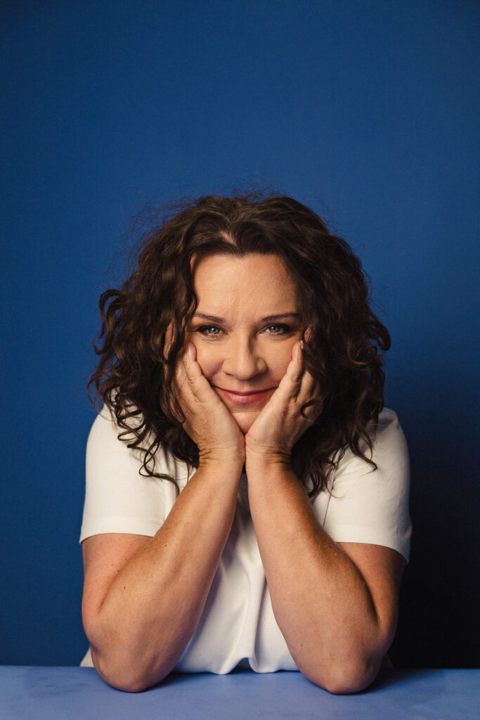 Zdjęcie portretowe aktorki Beaty Schimscheiner, delikatny uśmiech, ręce podpierają brodę.