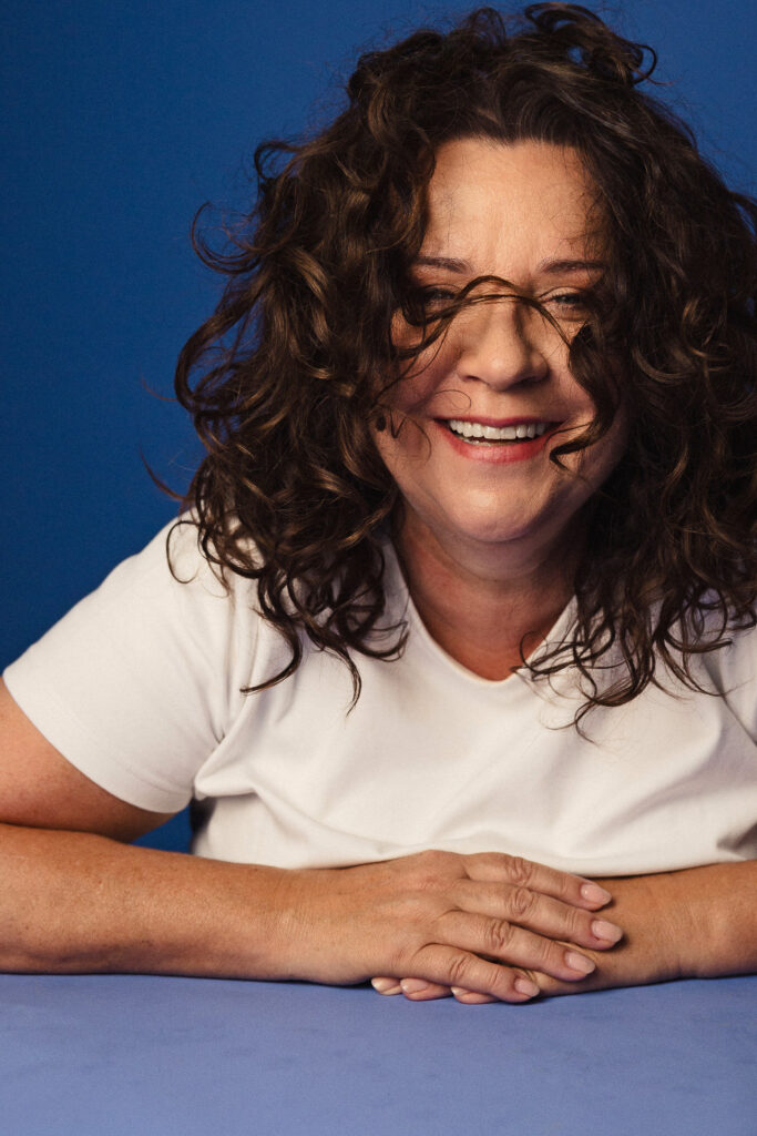 Zdjęcie portretowe aktorki Beaty Schimscheiner, delikatny uśmiech, włosy padające w nieładzie na twarz, ręce przed sylwetką.