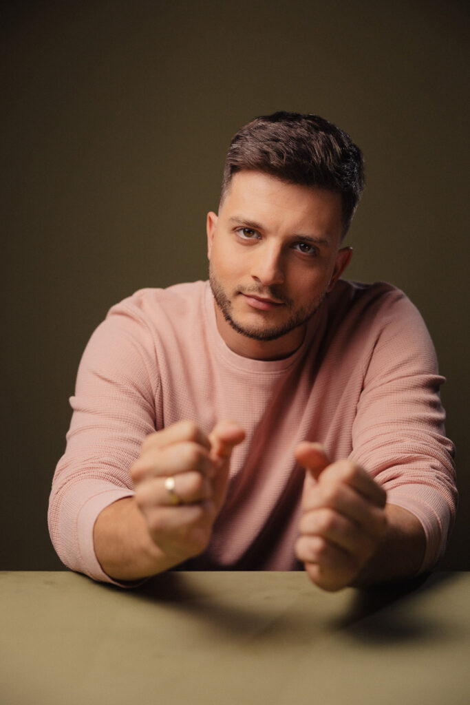 Zdjęcie portretowe aktora Roberta Ratusznego, lekki uśmiech, ręce wyciągnięte przed siebie.