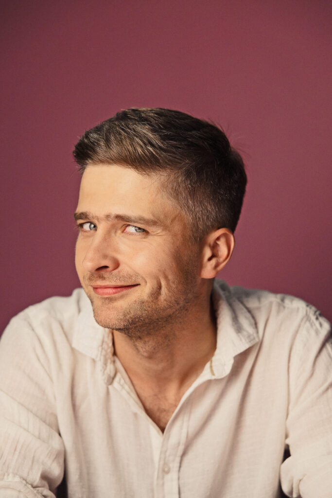 Zdjęcie półprofil portretowe aktora Karola Polaka, delikatny uśmiech, bliski kadr.