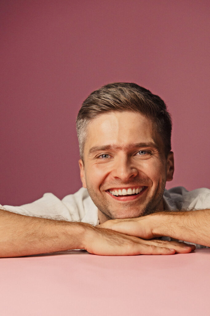 Zdjęcie portretowe aktora Karola Polaka, roześmiana twarz, bliski kadr, ręce pod brodą.