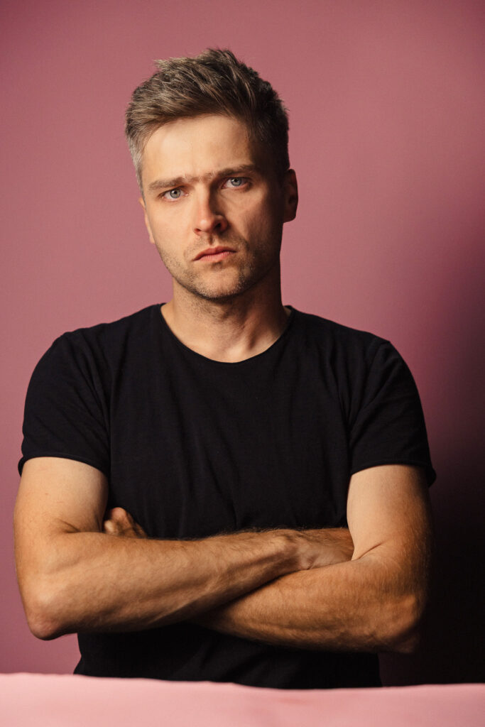 Zdjęcie portretowe aktora Karola Polaka, poważna twarz, bliski kadr., ręce skrzyżowana przed sobą