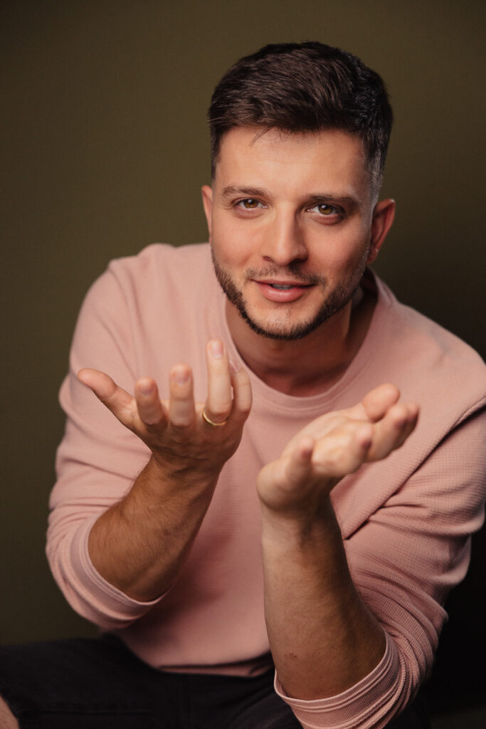 Zdjęcie portretowe aktora Roberta Ratusznego, dynamiczne, lekki uśmiech, ręce wyciągnięte do obiektywu.