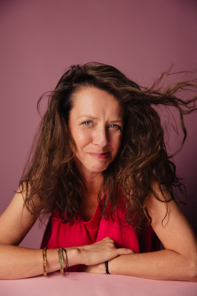 Zdjęcie portretowe aktorki Iwony Sitkowskiej, półuśmiech,oddmuchiwanie włosów z twarzy, ręce skrzyżowane przed sobą.