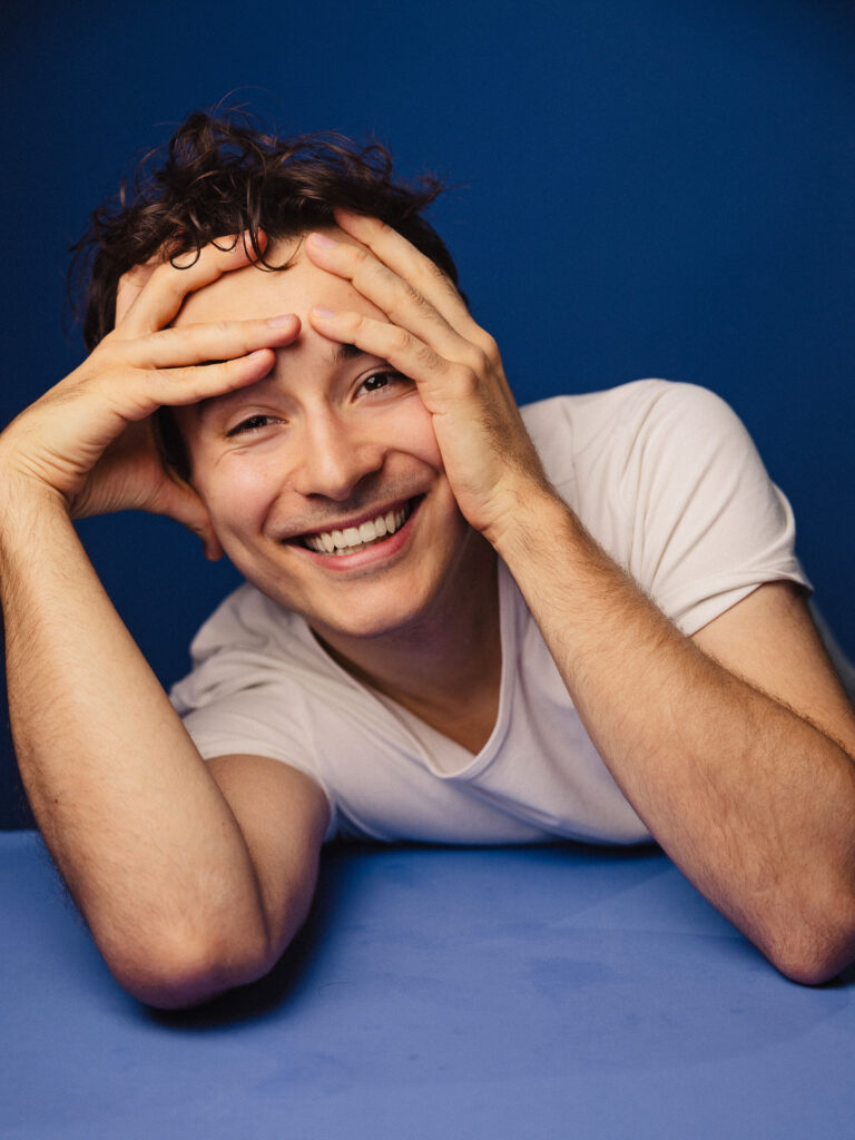 Zdjęcie portretowe aktora Antoniego Włosowicza, szeroki uśmiech, ręce na twarzy.