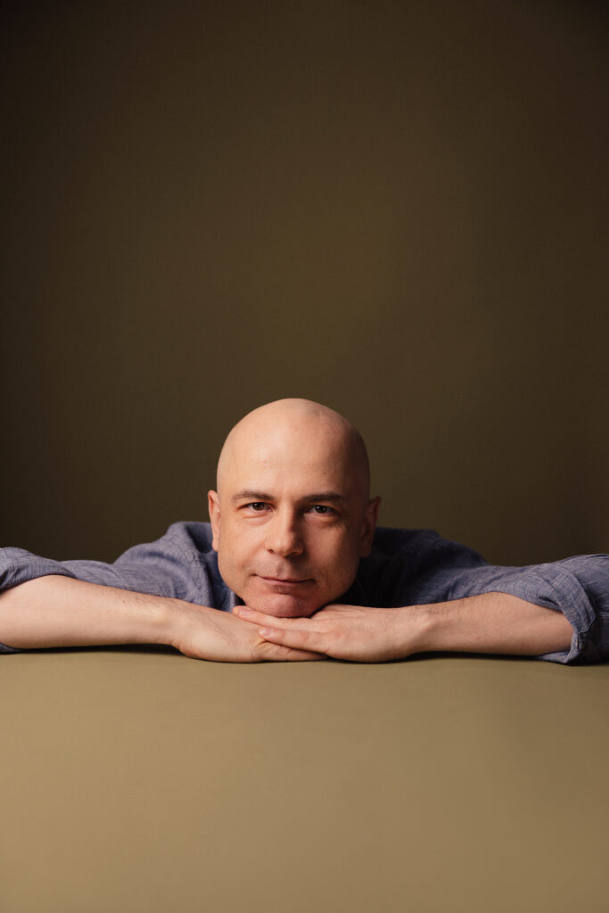 Zdjęcie portretowe aktora Macieja Namysło, lekki uśmiech, daleki kadr, ręce pod brodą.