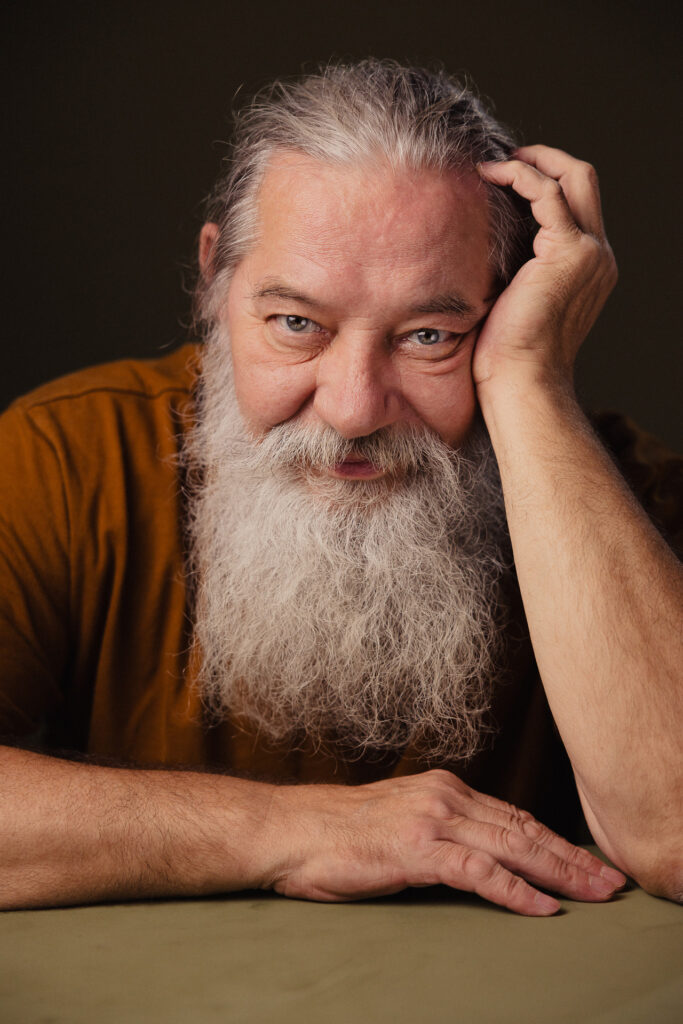 Zdjęcie portretowe aktora Kajetana Wolniewicza, uśmiechnięta twarz, bliski kadr, ręka opiera się o twarz druga przed osobą.