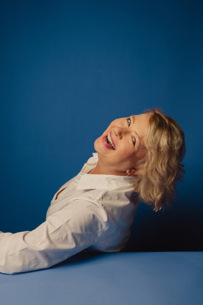 Zdjęcie półprofil portretowe aktorki Małgorzaty Kochan, dynamiczne, szeroki uśmiech, ręce przed sobą.