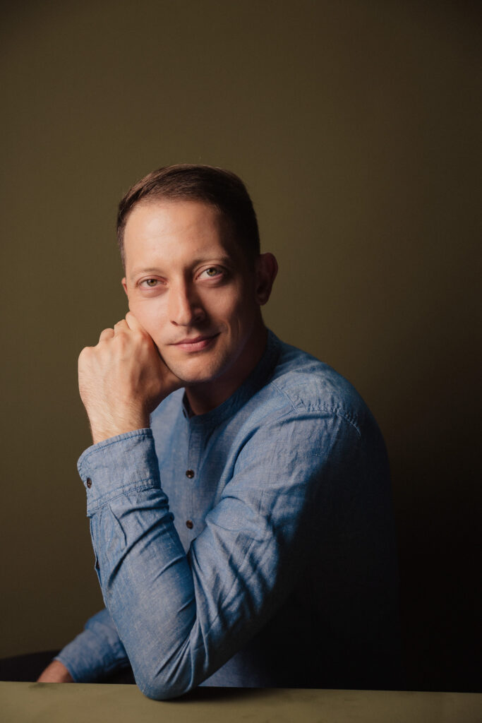 Zdjęcie profilowe portretowe aktora Wojciecha Lato, lekki uśmiech, ręka oparta o twarz.