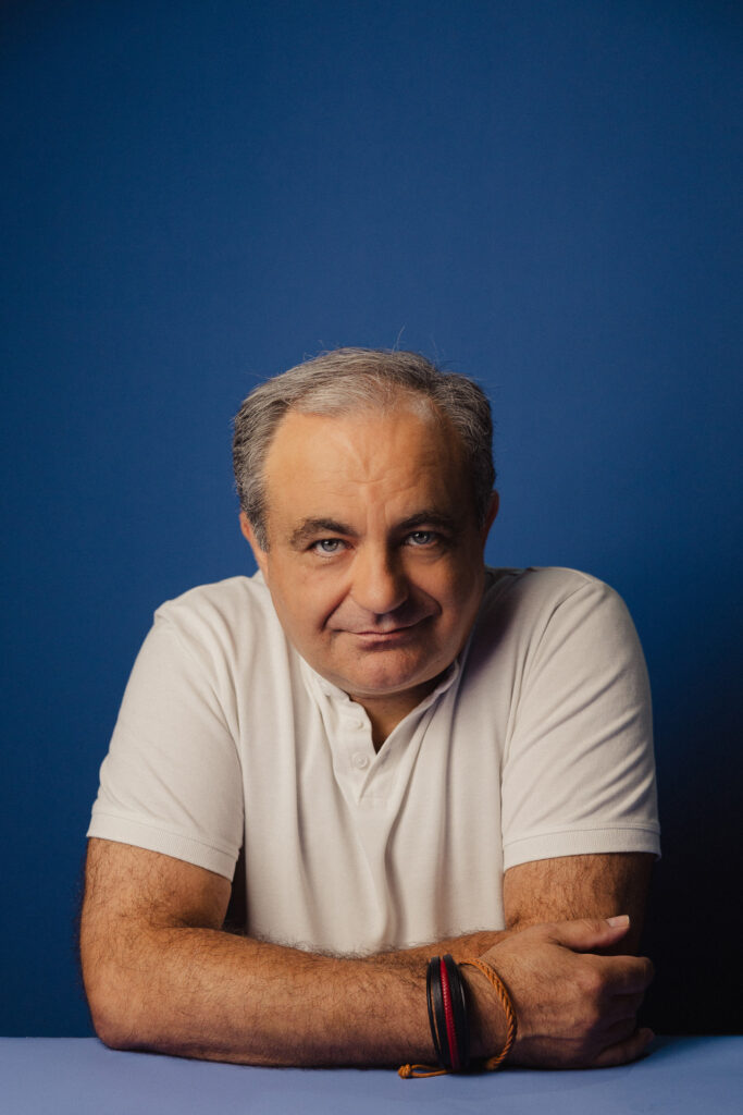 Zdjęcie profilowe portretowe aktora Tadeusza Łomnickiego, lekki uśmiech, ręce splecione przy sylwetce..