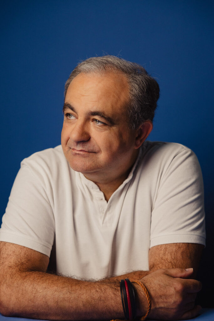 Zdjęcie półprofil profilowe portretowe aktora Tadeusza Łomnickiego, lekki uśmiech, ręce splecione przy sylwetce..