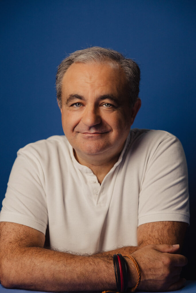 Zdjęcie półprofil profilowe portretowe aktora Tadeusza Łomnickiego, lekki uśmiech, ręce splecione przy sylwetce..