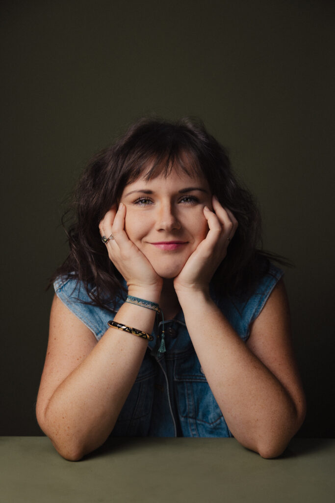 Zdjęcie portretowe aktorki Weroniki Kowalskiej, lekki uśmiech, bliski kadr, ręce otulają i podpierają twarz.