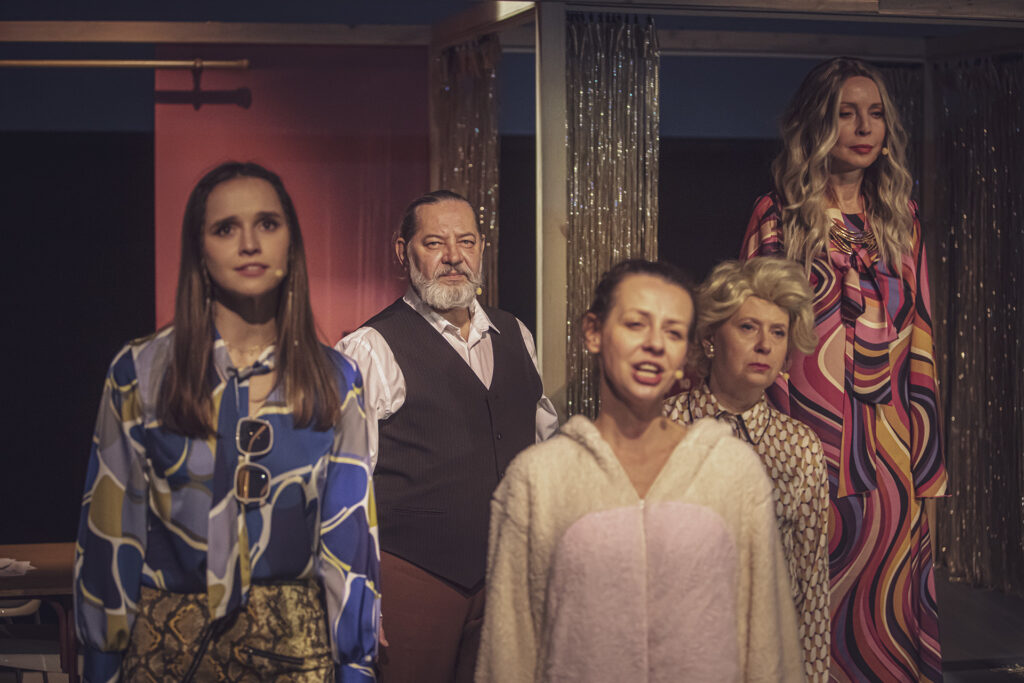 Zdjęcie ze spektaklu Aktorzy prowincjonalni. Cztery aktorki i aktor stoją blisko siebie i patrzą przed siebie. Aktorka w jednoczęściowej piżamie otwiera usta. Pozostałe aktorki mają wzorzyste stroje, a aktor ma koszulę i kamizelkę.