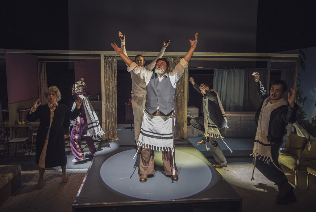 Zdjęcie ze spektaklu Aktorzy prowincjonalni. Na małej scenie stoi starszy aktor, za nim jest młody aktor. Aktorzy mają podniesione ręce i śpiewają. Wokół nich chodzą dwie aktorki i dwóch aktorów. Na kostiumy mają narzucone płachty z frędzlami.