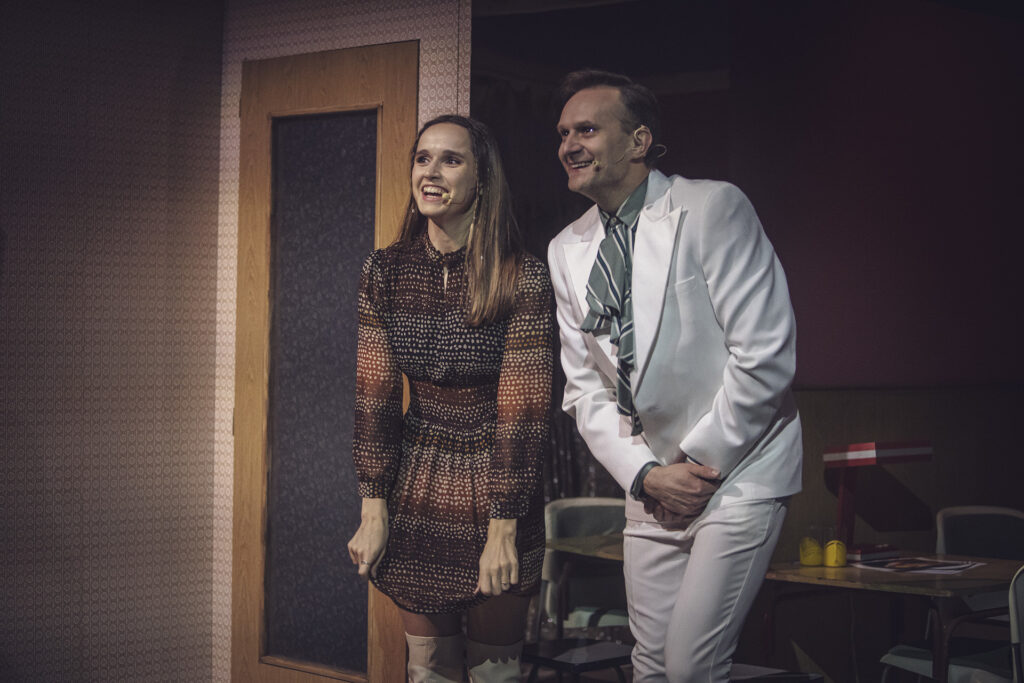 Zdjęcie ze spektaklu Aktorzy prowincjonalni. Aktorka i aktor stoją uśmiechnięci. Aktorka trzyma brzeg swojej sukienki, a aktor w garniturze trzyma złączone dłonie przed sobą.