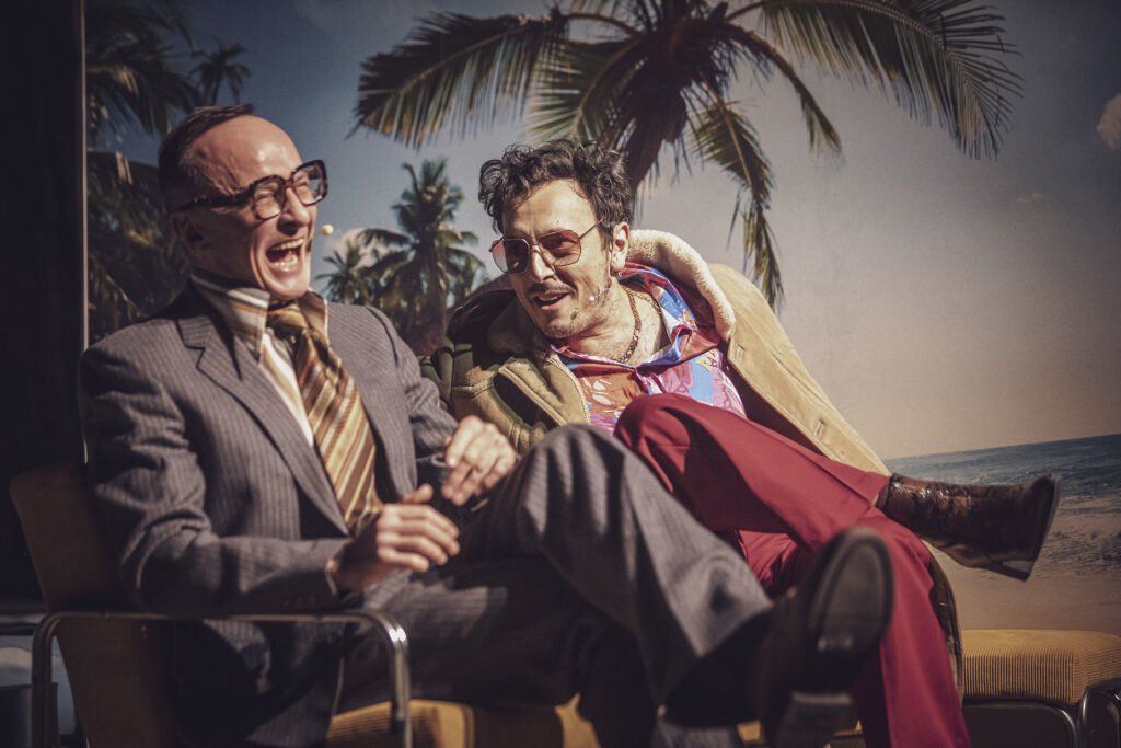 Zdjęcie ze spektaklu Aktorzy prowincjonalni. Na fotelach siedzą dwaj aktorzy i się śmieją. Jeden ma garnitur i okulary, a drugi kolorowe ubrania, kożuch i okulary przeciwsłoneczne. Za nimi na ścianie jest fototapeta z palmą i morzem.