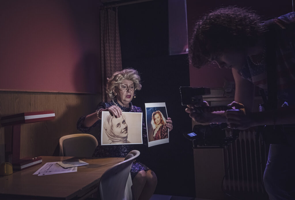 Zdjęcie ze spektaklu Aktorzy prowincjonalni. Starsza aktorka w tapirowanych włosach i okularach siedzi przy stole. Trzyma dwa zdjęcia młodych kobiet. Przed kobietą stoi młody mężczyzna z kamerą i ją nagrywa.