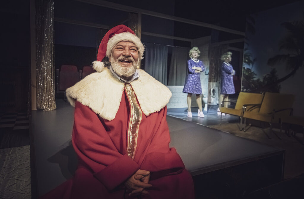 Zdjęcie ze spektaklu Aktorzy prowincjonalni. Z przodu jest starszy aktor w stroju świętego Mikołaja. W tle przy lustrze stoi aktorka w tapirowanych włosach. Aktorka ma krótką sukienkę i buty na obcasie. Aktorka krzyżuje ręce i patrzy na mężczyznę.