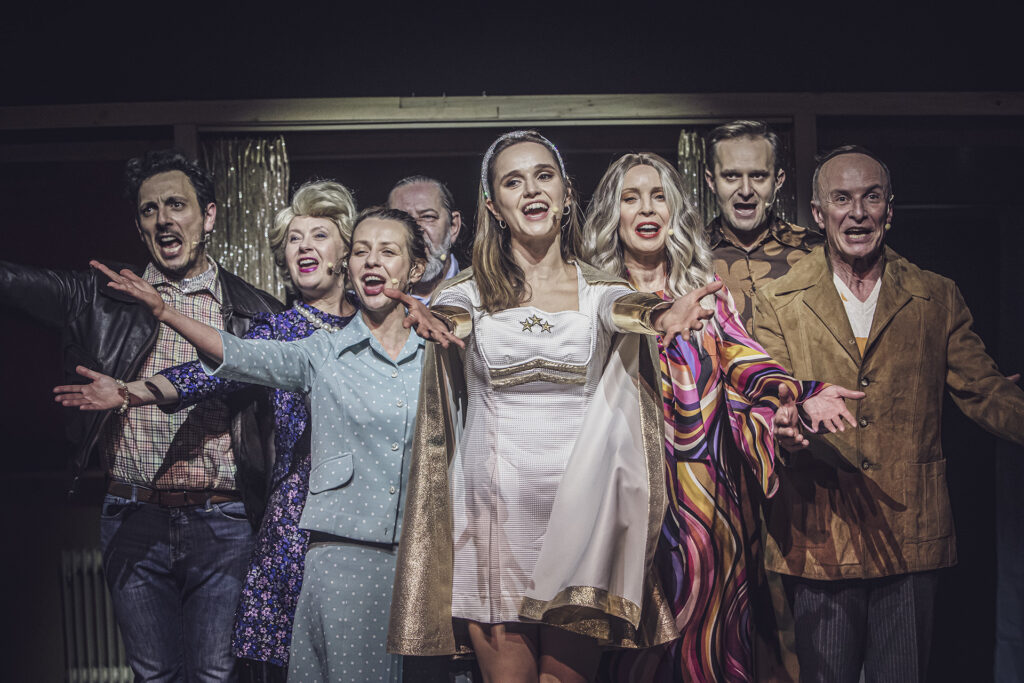 Zdjęcie ze spektaklu Aktorzy prowincjonalni. Cztery aktorki i czterech aktorów w różnym wieku stoi obok siebie. Aktorki stoją na pierwszym planie. Wszyscy mają kolorowe stroje w hollywoodzkim stylu. Rozkładają szeroko ręce i śpiewają.