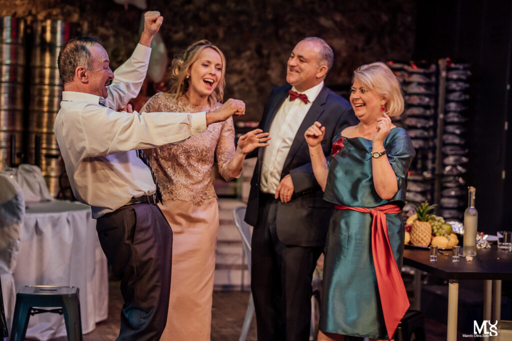 Zdjęcie ze spektaklu Wstyd. Dwie kobiety i dwaj mężczyźni w średnim wieku stoją obok siebie na zapleczu sali weselnej. Śmieją się do siebie. Kobiety są w sukniach balowych, a mężczyźni w garniturach. Za nimi jest stół z owocami i wódką.