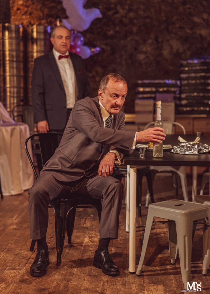 Zdjęcie ze spektaklu Wstyd. Na pierwszym planie aktor z krawatem siedzi przy stole i trzyma butelkę wódki. Drugi aktor z muchą stoi za nim.