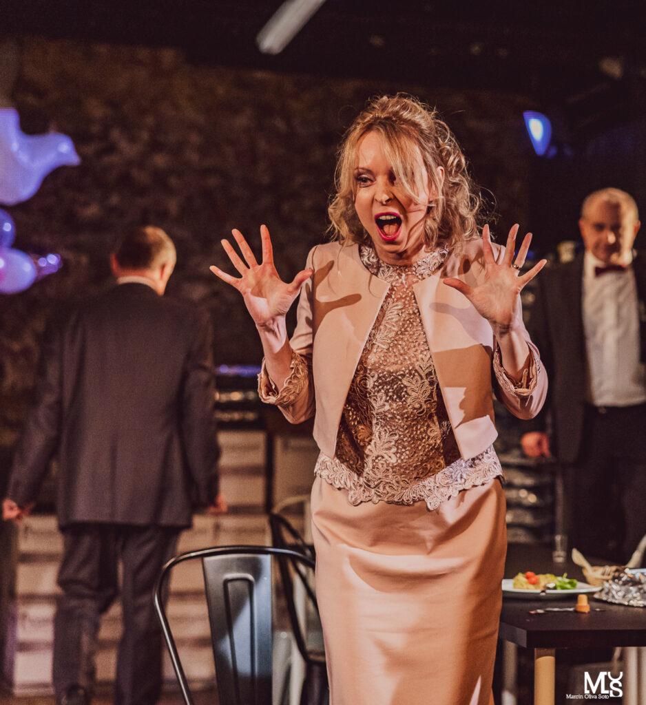 Zdjęcie ze spektaklu Wstyd. Na pierwszym planie aktorka z kręconymi włosami w sukni balowej otwiera usta i podnosi ręce do góry. Jest zdziwiona. W tle stoją dwaj aktorzy.