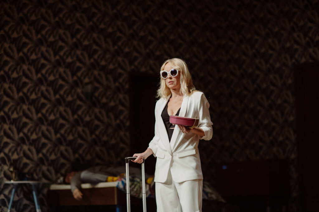 Zdjęcie ze spektaklu Akt równoległy. Aktorka w średnim wieku. Ma długie włosy i okulary przeciwsłoneczne. Na sobie ma garnitur i bluzkę z dekoltem. W jednej ręce trzyma rączkę walizki. A w drugiej pudełko. W tle na łóżku leży mężczyzna.