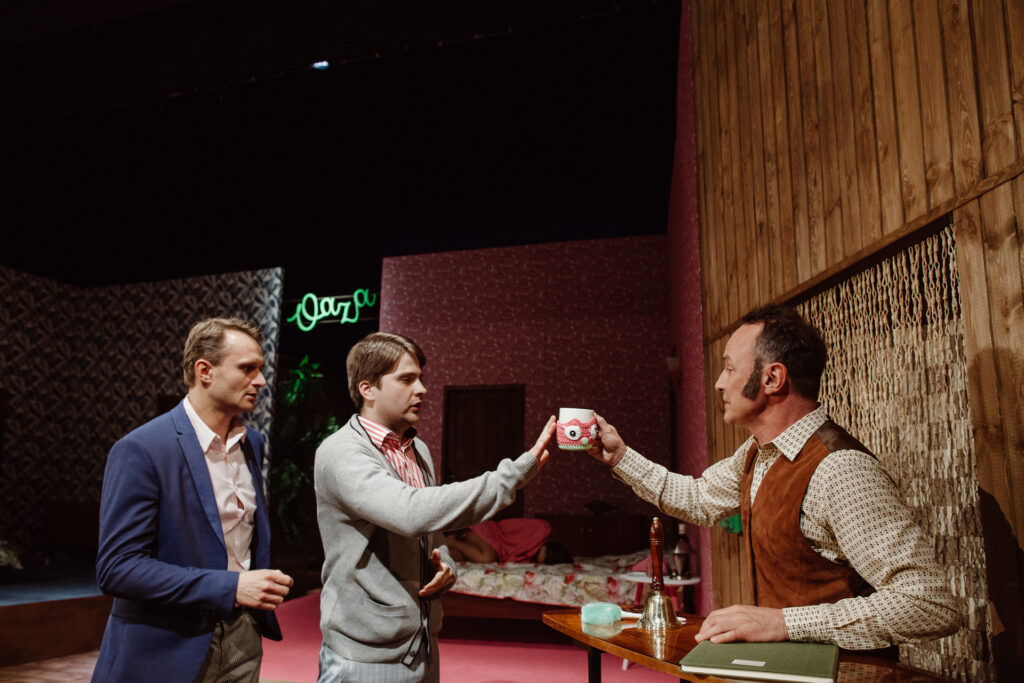 Zdjęcie ze spektaklu Akt równoległy. Scenografia przedstawia pensjonat. Jeden aktor stoi za ladą. Ma kamizelkę, na twarzy ma baki, trzyma kubek z oczami sowy. Po drugiej stronie stoi dwóch młodych aktorów. Na ścianie jest napis Oaza.