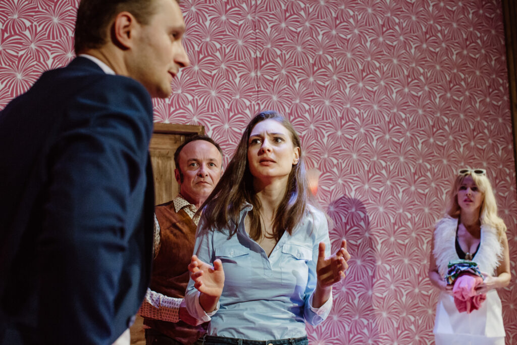 Zdjęcie ze spektaklu Akt równoległy. Na pierwszym planie młoda aktorka patrzy na aktora w marynarce. Aktor odwraca głowę. Z tyłu stoi aktor z bakami i aktorka w garniturze. Wszyscy są zdenerwowani.