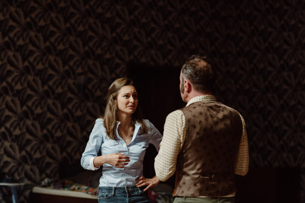 Zdjęcie ze spektaklu Akt równoległy. Młoda aktorka patrzy na aktora w średnim wieku. Aktor stoi tyłem. Aktorka jedną dłoń trzyma na biodrze, a drugą na wysokości mostka.