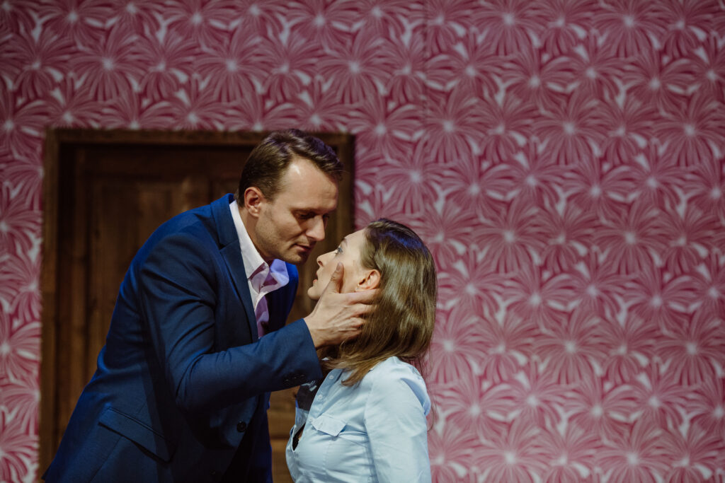 Zdjęcie ze spektaklu Akt równoległy. Młody aktor i młoda aktorka patrzą na siebie. Są elegancko ubrani. Aktor dłonią dotyka twarzy aktorki. W tle jest kolorowa tapeta na ścianie.