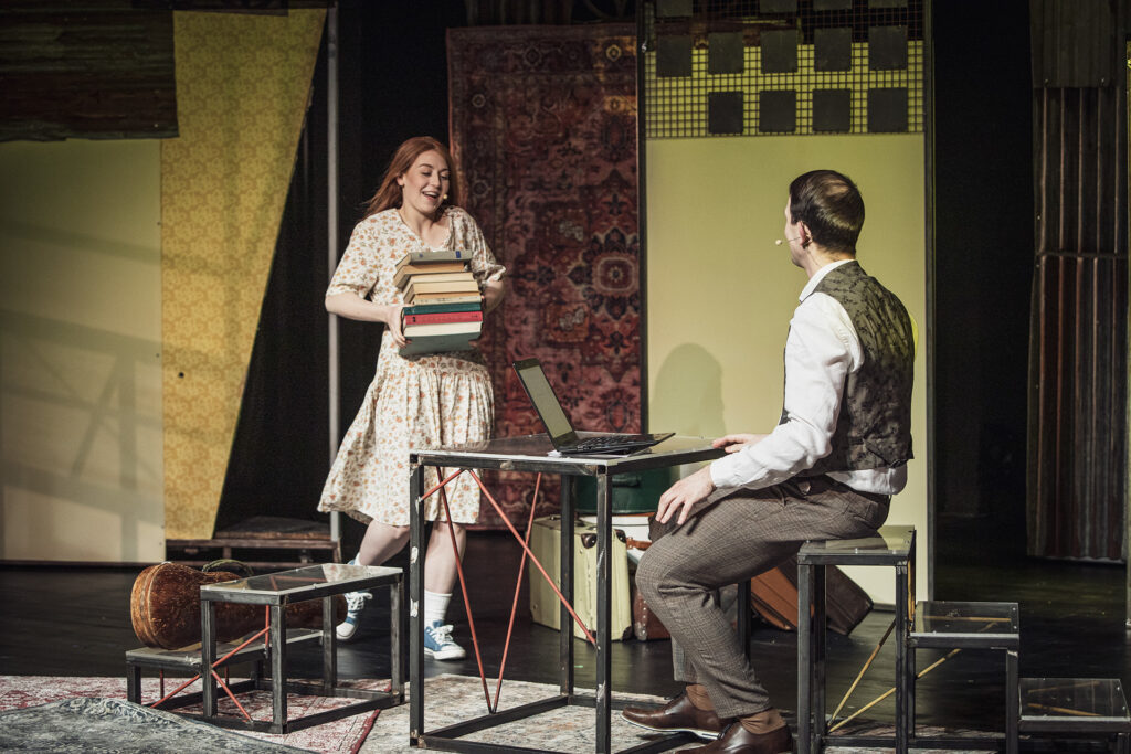 Zdjęcie ze spektaklu Mieszkańcy sąsiednich domów. Aktor w eleganckim stroju siedzi przy stole przed laptopem. Aktorka w sukience w kwiaty niesie stos książek. W tle są walizki i ściany bloków mieszkalnych z papieru.