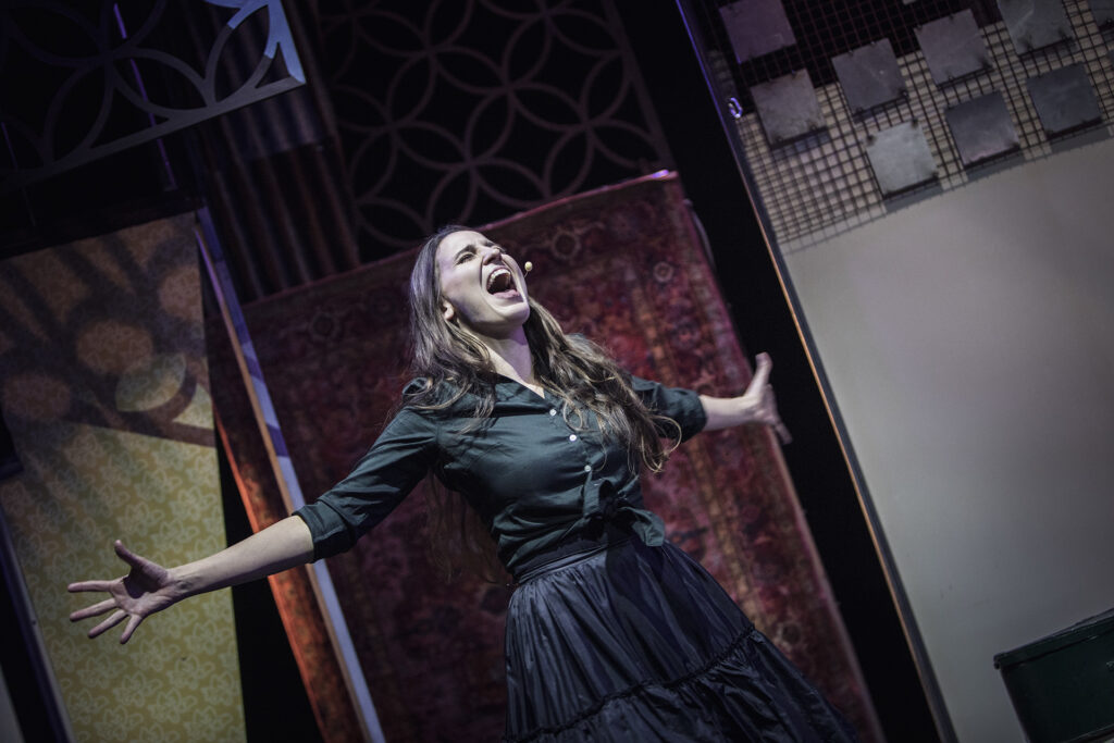 Zdjęcie ze spektaklu Mieszkańcy sąsiednich domów. Aktorka rozkłada ręce, zamyka oczy i otwiera usta jak do krzyku. W tle są bloki mieszkalne z papieru.