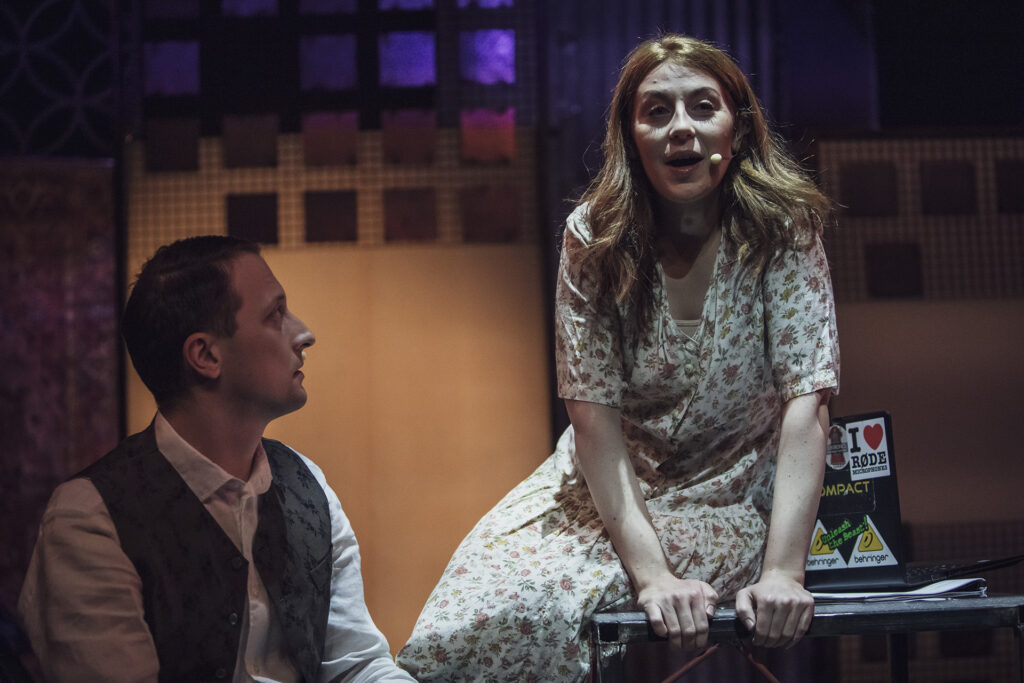 Zdjęcie ze spektaklu Mieszkańcy sąsiednich domów. Aktorka w sukni w kwiaty siedzi na stole i otwiera usta. Obok siedzi aktor w koszuli i kamizelce. W tle są bloki mieszkalne z papieru.