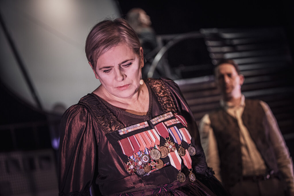 Zdjęcie ze spektaklu Mieszkańcy sąsiednich domów. Zbliżenie. Na pierwszym planie aktorka spuszcza wzrok. Ma do sukni przypięte różne ordery. W tle stoi aktor w kamizelce.