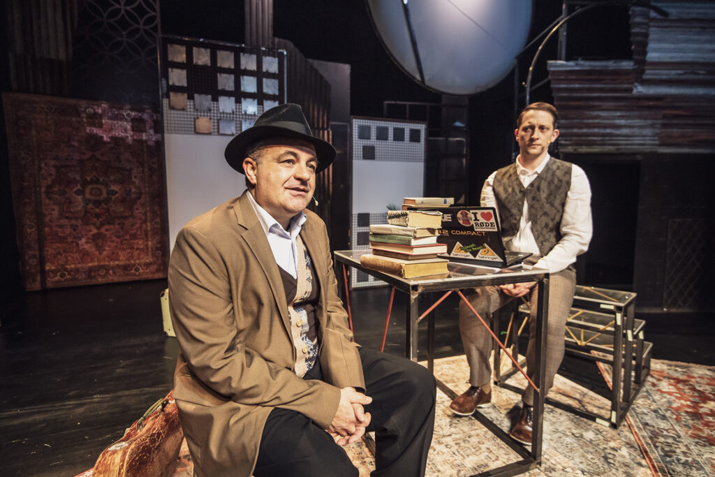 Zdjęcie ze spektaklu Mieszkańcy sąsiednich domów. Przy stole siedzi starszy aktor w płaszczu i kapeluszu oraz młody aktor w eleganckim stroju. Młody aktor patrzy na starszego. Na stole jest laptop i książki. W tle są bloki mieszkalne z papieru.