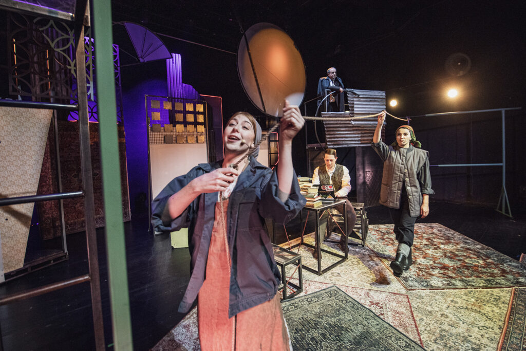 Zdjęcie ze spektaklu Mieszkańcy sąsiednich domów. Dwie aktorki w robotniczych strojach trzymają sznurek na pranie. Z tyłu aktor w eleganckim stroju siedzi przy stole z laptopem. W tle starszy aktor stoi na wysokim podeście i opiera się o barierkę.