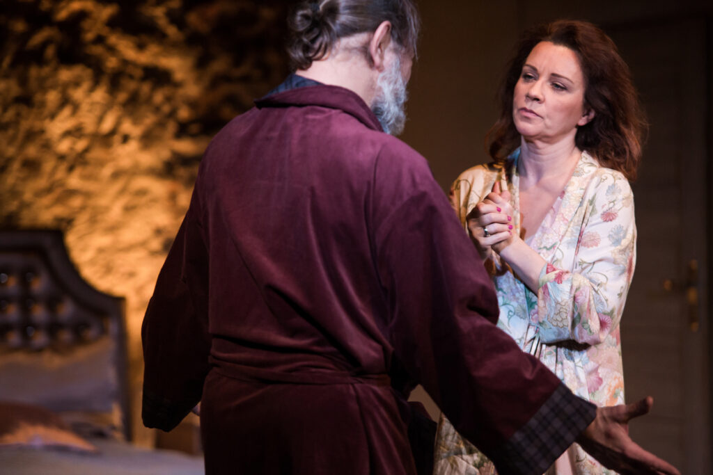 Zdjęcie ze spektaklu Via-gra. Aktorka stoi naprzeciwko aktora. Aktorka ma szlafrok i obrączkę na dłoni. Aktor rozkłada ręce. Ma szlafrok i włosy związane w koka.