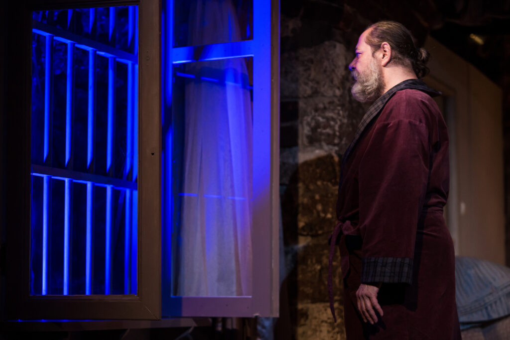 Zdjęcie ze spektaklu Via-gra. Aktor stoi bokiem przed otwartym oknem. Aktor ma szlafrok, długą siwą brodę i włosy związane w koka.