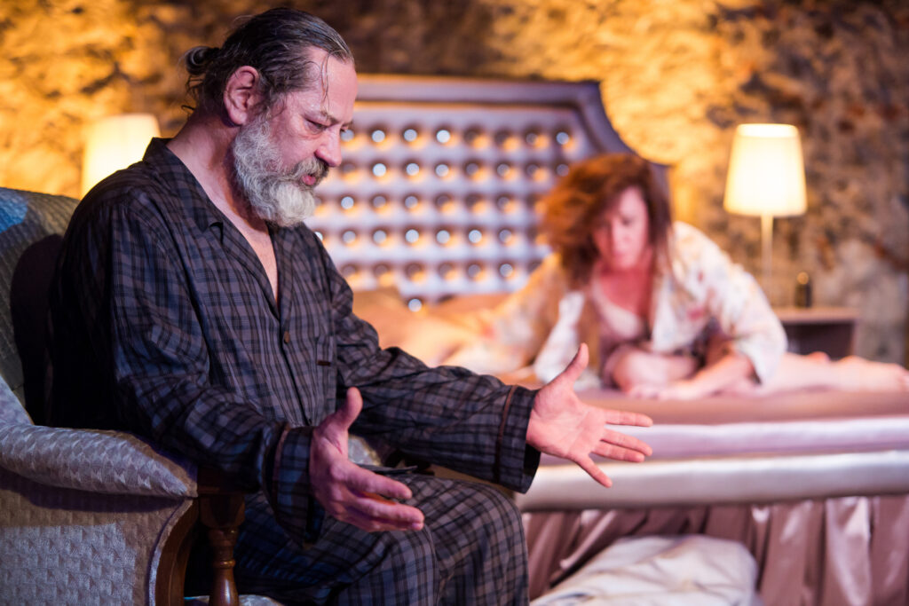 Zdjęcie ze spektaklu Via-gra. Na pierwszym planie aktor siedzi bokiem na fotelu i rozkłada ręce. Aktor ma piżamę w kratkę, długą siwą brodę i włosy związane w koka. W tle aktorka w szlafroku siedzi na łóżku i pochyla się do przodu.