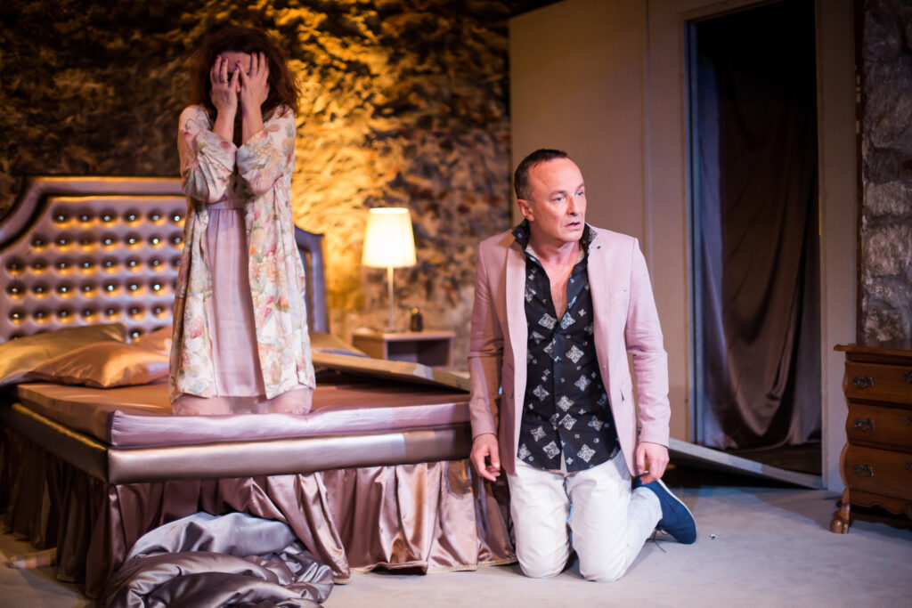 Zdjęcie ze spektaklu Via-gra. Aktorka w szlafroku klęczy na łóżku i zakrywa twarz dłońmi. Przed łóżkiem klęczy mężczyzna w garniturze, patrzy w bok, jest zaskoczony.