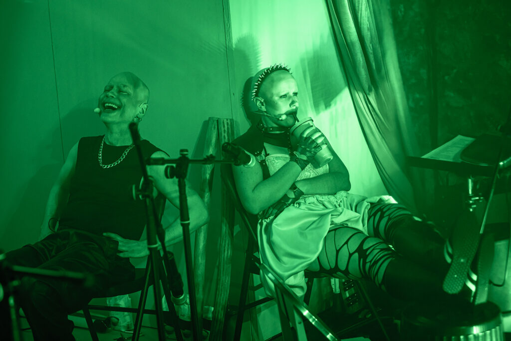 Zdjęcie ze spektaklu Saga rodu Szyców. Zdjęcie zabarwione na zielono. Łysy aktor i łysa aktorka siedzą na krzesłach przy ścianie. Aktorka opiera nogi na bębnie i pije z kubka ze słomką. Aktor zamyka oczy i się śmieje.