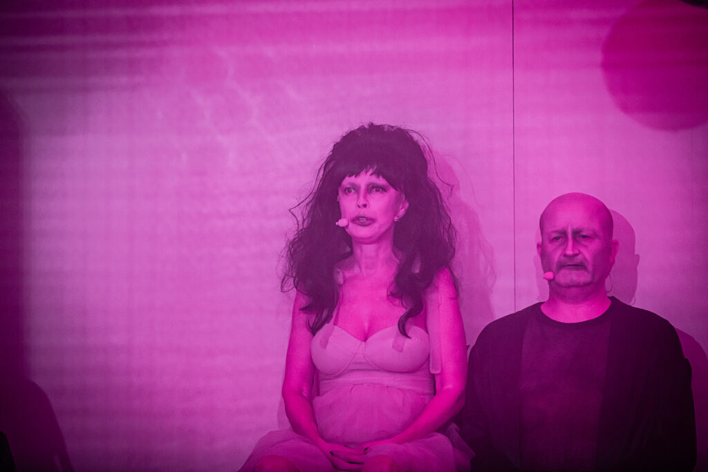 Zdjęcie ze spektaklu Saga rodu Szyców. Zdjęcie zabarwione na różowo. Zbliżenie. Aktorka i aktor siedzą przy ścianie. Aktorka ma tiulową sukienkę, perukę, bladą twarz i rozmazany makijaż. Aktor jest łysy, ma ciemny strój i pobrudzoną twarz.